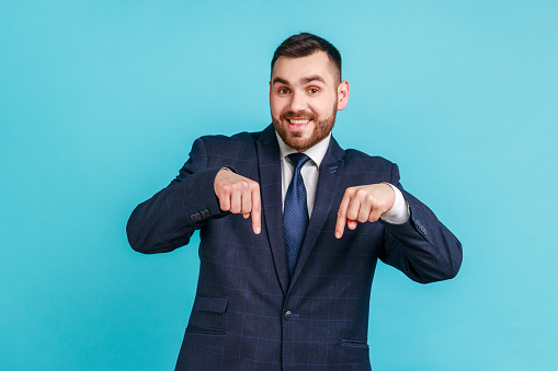 Portrait of attractive bearded businessman wearing official style suit pointing down with fore fingers, looking at camera with toothy smile. Indoor studio shot isolated on blue background.