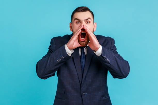 Portrait of hysterical young adult man wearing official style suit loudly yelling in panic, holding hands near his mouth, teenager protest. Portrait of hysterical young adult man wearing official style suit loudly yelling in panic, holding hands near his mouth, teenager protest. Indoor studio shot isolated on blue background. Phobia stock pictures, royalty-free photos & images