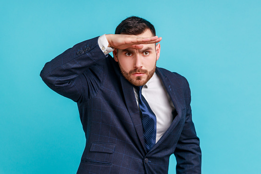 Far future. Portrait of handsome man wearing official style suit looking into distance, holding hand over eyes and watching horizon with attentive view. Indoor studio shot isolated on blue background.