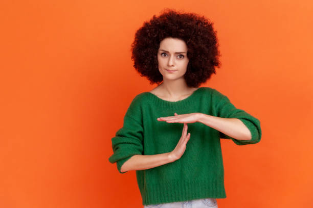Young adult woman with Afro hairstyle wearing green casual style sweater showing time out hand gesture, looking at camera, worried about deadline. Young adult woman with Afro hairstyle wearing green casual style sweater showing time out hand gesture, looking at camera, worried about deadline. Indoor studio shot isolated on orange background. time out signal stock pictures, royalty-free photos & images