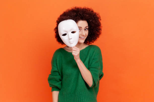 Woman with Afro hairstyle in green sweater removing white mask from face showing his smiling expression, good mood, pretending to be another person. Woman with Afro hairstyle in green sweater removing white mask from face showing his smiling expression, good mood, pretending to be another person. Indoor studio shot isolated on orange background. hypocrisy stock pictures, royalty-free photos & images