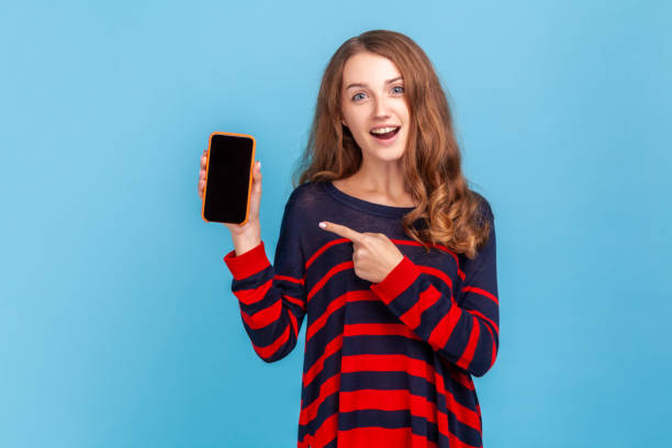 istock Woman pointing at cell phone with blank black screen for advertisement, having surprised expression. 1392625230
