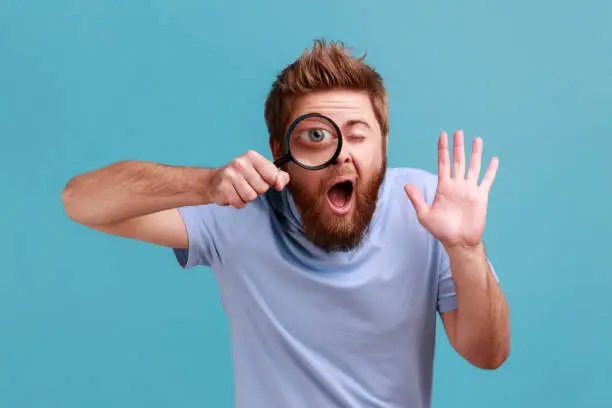 Photo of Man holding magnifying glass and looking at camera with big zoom eye, waving hand, saying hi.