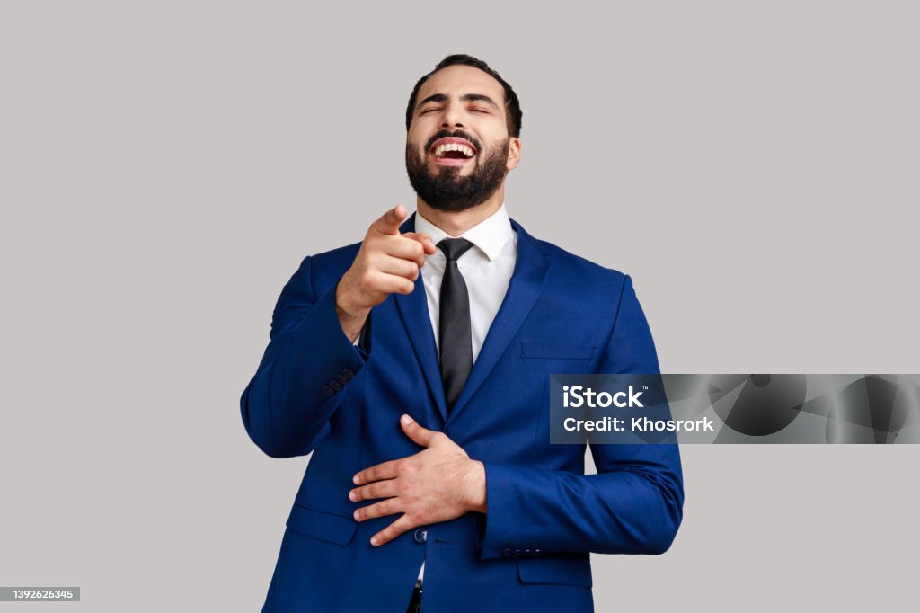 Bearded man laughing out loud holding belly and pointing finger on you, mockery. Positive excited bearded man laughing out loud holding belly and pointing finger on you, mockery, wearing official style suit. Indoor studio shot isolated on gray background. Sneering Stock Photo