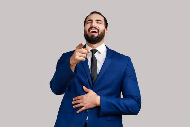 Positive excited bearded man laughing out loud holding belly and pointing finger on you, mockery, wearing official style suit. Indoor studio shot isolated on gray background.