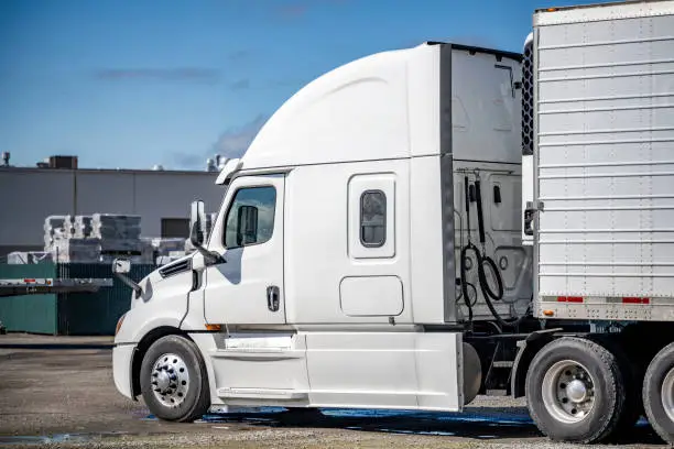 Photo of Popular bonnet big rig semi truck in white color with refrigerator semi trailer waiting for the next load standing on the industrial warehouse parking lot