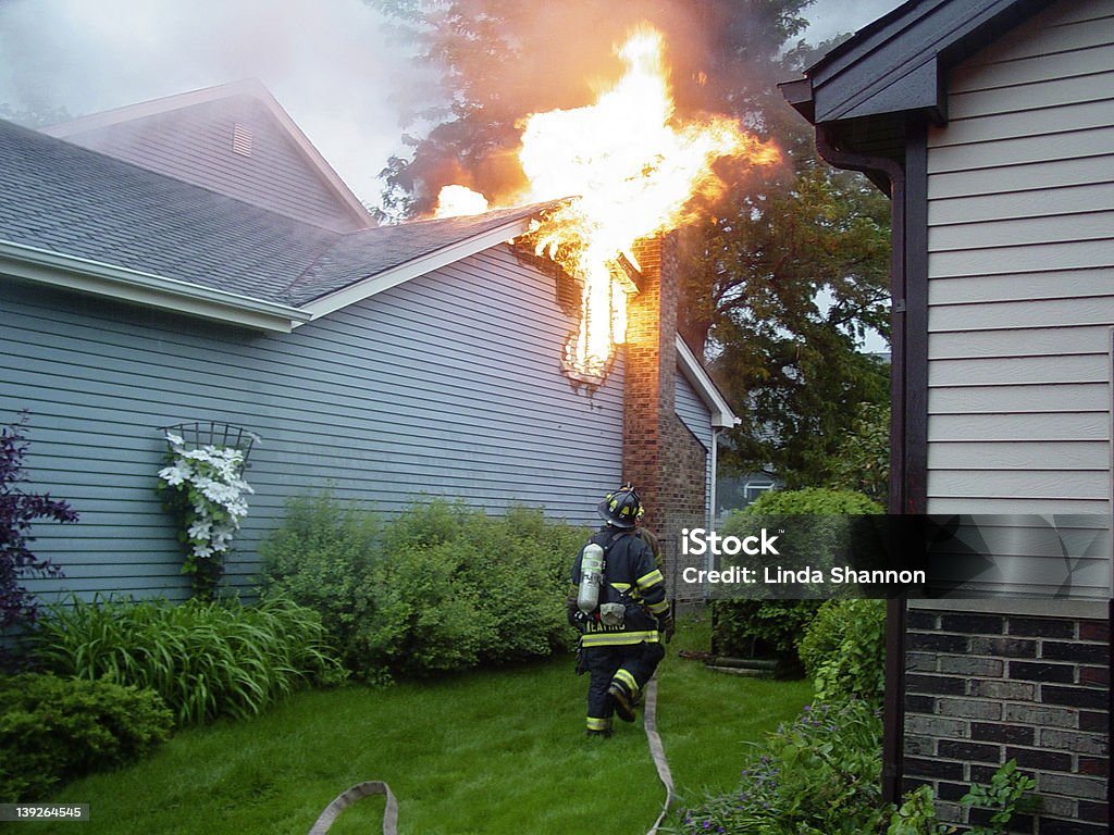 Fighting a House Fire The roof of a house is burning, while a firefighter rushes with his hose to put out the blaze. Fire - Natural Phenomenon Stock Photo