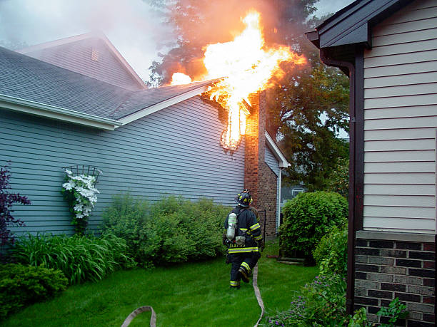 Fighting a House Fire The roof of a house is burning, while a firefighter rushes with his hose to put out the blaze. natural phenomenon stock pictures, royalty-free photos & images