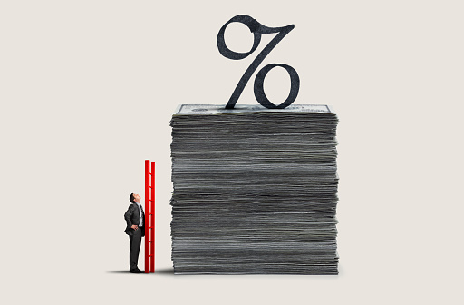 A man stands with a red ladder as he looks up at a tall stack of money topped with an interest rate symbol.
