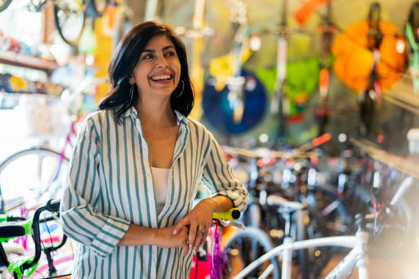 Latin american adult woman smiling while leaning on bicycle at store stock photo