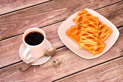 Puff pastries with apricot jam on a plate and a cup of hot black coffee on a wooden table. Close-up.