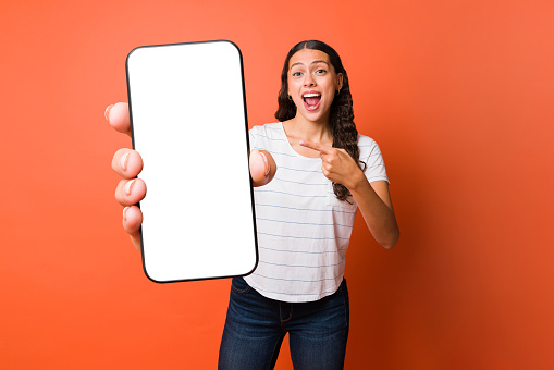 Surprised young woman pointing to the white screen of her smartphone while using online mobile apps