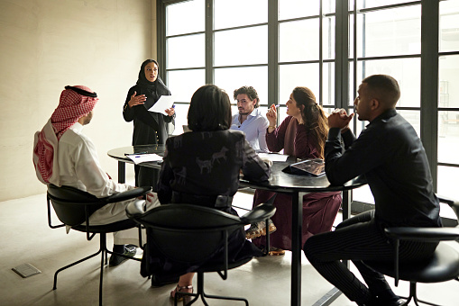 Black and Middle Eastern professionals sitting around conference table in modern office, listening to project manager describe plans, and asking questions.