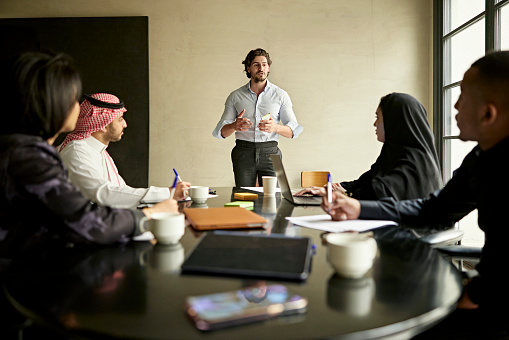 Black and Middle Eastern associates in traditional and western attire sitting around conference table listening to project manager’s plans for development.