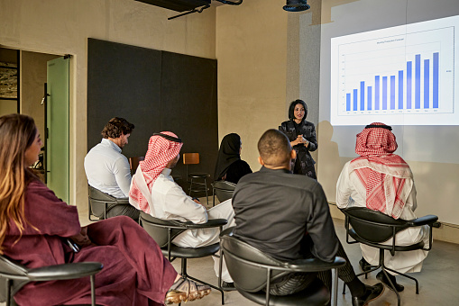 Middle Eastern men and women in traditional and western attire sitting in audience, looking at bar graph on projection screen, and listening to project update.