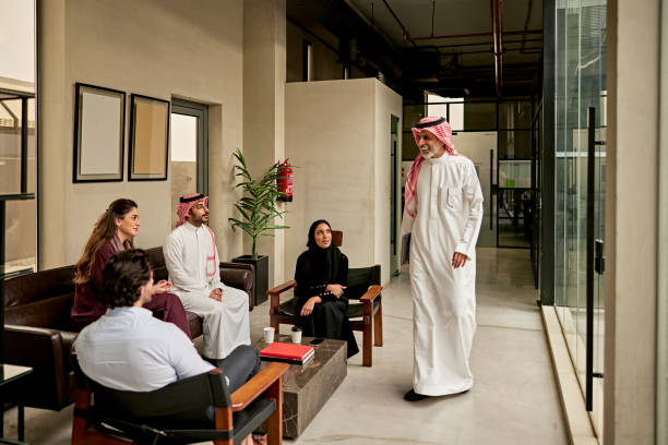 Middle Eastern executive arriving for informal meeting Full length front view of mature Saudi businessman in traditional attire smiling at project team members gathered for quick update in modern office environment. religious dress stock pictures, royalty-free photos & images