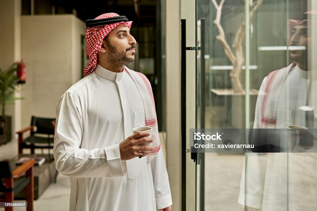 Indoor portrait of late 20s Riyadh businessman with coffee Waist-up view of Middle Eastern man in dish dash, kaffiyeh, and agal taking a break for coffee and a moment of contemplation while looking through window. Saudi Arabia Stock Photo