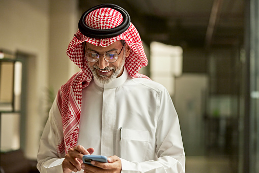 Waist-up front view of bearded Middle Eastern man in late 40s wearing traditional attire and showing positive emotion while looking at portable device.