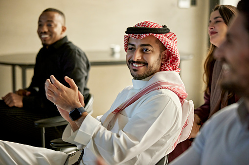 Candid portrait of Middle Eastern man in dish dash, kaffiyeh, and agal sitting among associates, smiling, and clapping after presentation.