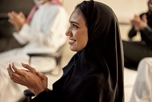 Profile view of young Middle Eastern executive in black abaya and headscarf smiling and clapping at conclusion of presentation.