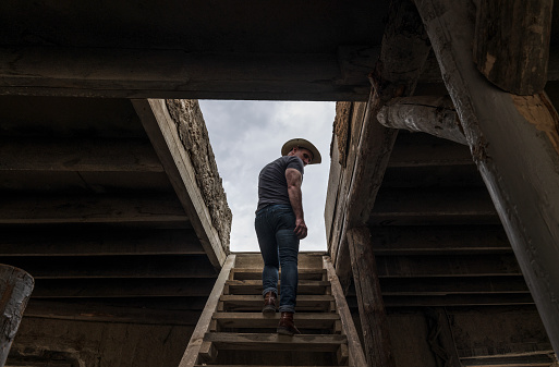 Rear view of adult man in cowboy hat climbing up wooden stairs, indoor toward rooftop