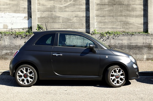 Udine, Italy. April 16, 2022. Matt black new Fiat 500 car with grunge concrete wall on background. Side view.
