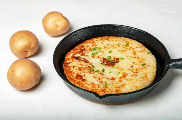 traditional Swiss dish is a potato pancake in a frying pan, decorated with green onions. whole tubers of unpeeled potatoes lie nearby. side view.