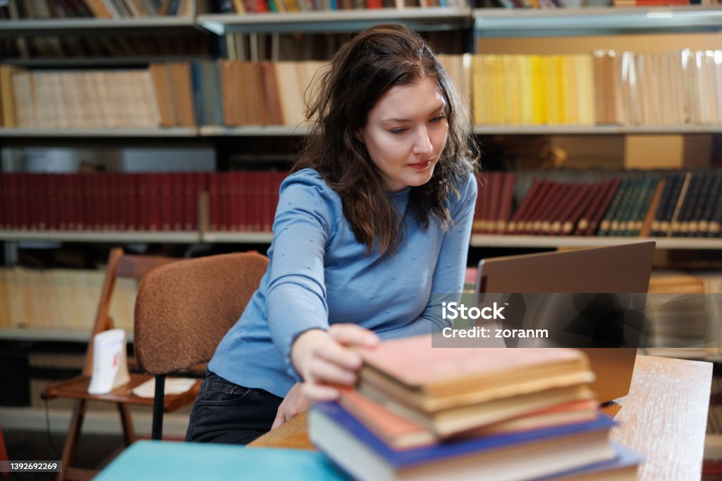 Beautiful post-grad using laptop and picking up book from stack on the desk in library Beautiful university student using laptop and picking up book from stack on the desk in university library Laptop Stock Photo