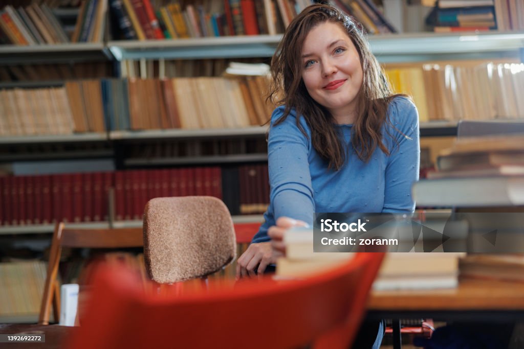 Beautiful post-grad sitting at the desk in university library with stack of books and smiling at camera Female university student sitting at the desk in the library with stack of books, looking at camera and smiling 30-34 Years Stock Photo