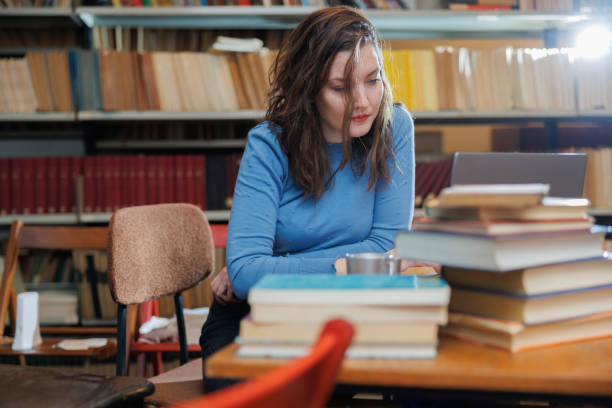 istock Beautiful university student sitting at the desk in university library with stack of books, looking tired 1392692294