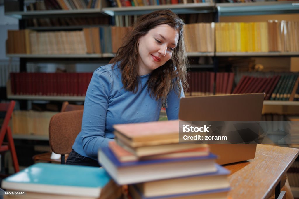 Female post-grad using laptop at the desk in university library and smiling Beautiful university student sitting in the library at a desk full of books and using laptop with a smile 30-34 Years Stock Photo