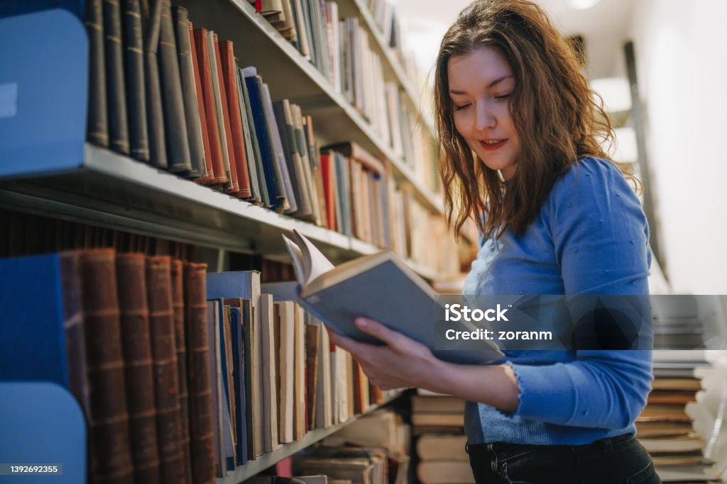 Post-grad holding a book with study material by the university library shelves Beautiful university student standing by the shelves in the library, holding and reviewing open book with study material 30-34 Years Stock Photo