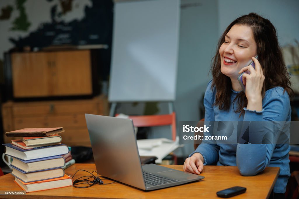 Beautiful post-grad using laptop and phone at the desk in the university office Beautiful university student using laptop and talking on the smart phone at the desk with stack of books in the office 30-34 Years Stock Photo