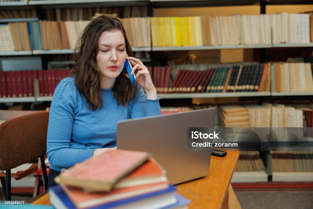 Beautiful post-grad using laptop and phone at the desk in the university library Beautiful university student using laptop for writing paper and talking on the smart phone at the desk in the library 30-34 Years Stock Photo