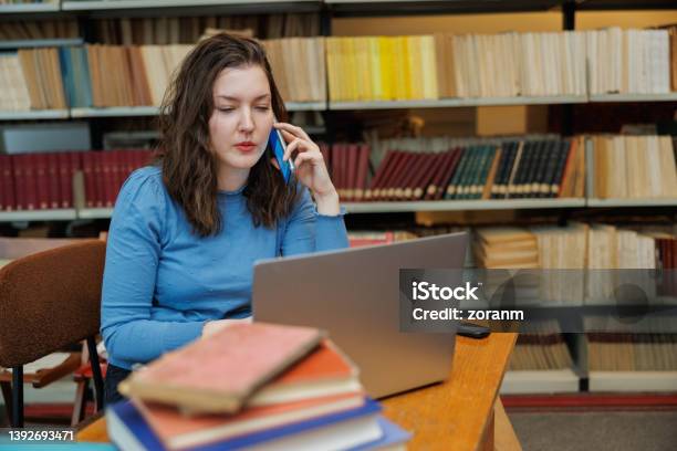 Beautiful Postgrad Using Laptop And Phone At The Desk In The University Library Stock Photo - Download Image Now