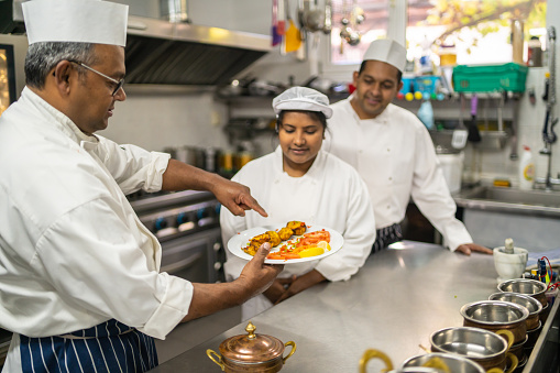 A chef is holding a plate of spicy chicken in one hand and pointing something out to his colleagues in an Indian restaurant kitchen