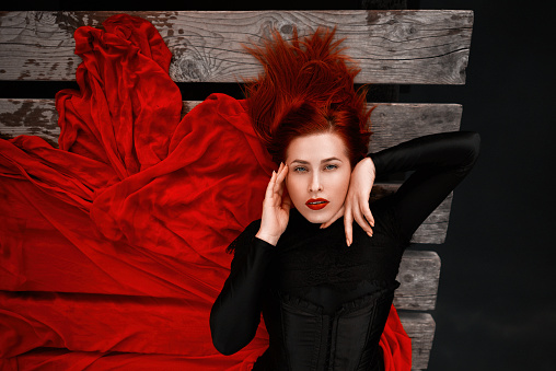 High angle view of woman with red hair and red lipstick wearing red skirt posing in fairytale concept  laying down on wooden pontoon and looking at camera