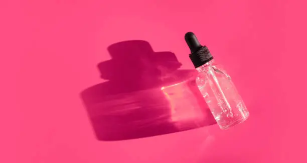 Transparent glass dropper bottle with air bubbles on bright pink purple background in the sunlight. Pipette with fluid hyaluronic acid, serum, retinol. Cosmetics, healthcare, beauty products concept