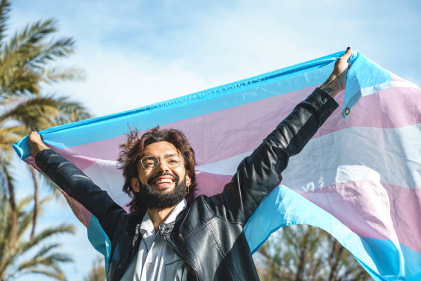 Happy man with transgender flag Low angle of cheerful young bearded man in casual clothes raising arms and smiling while standing on background of blue sky transgender stock pictures, royalty-free photos & images