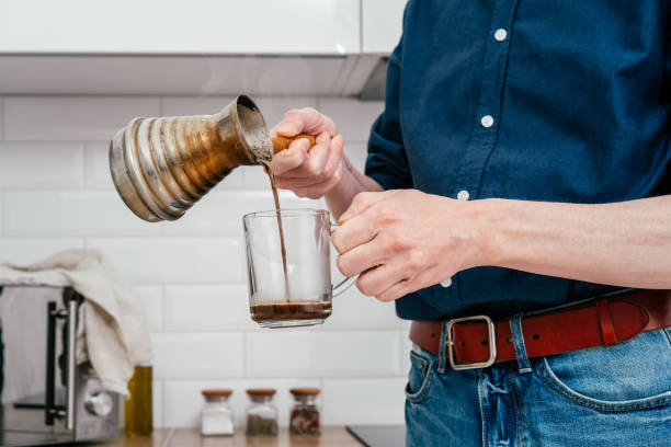 Hands of unrecognizable man in blue shirt pouring fresh coffee from copper cezve into glass cup at bright kitchen. Close up of hands of unrecognizable man in blue shirt pouring fresh brewed hot natural coffee from copper cezve with wooden handle into glass cup in white kitchen in morning. Making coffee at home. turkish coffee pot cezve stock pictures, royalty-free photos & images