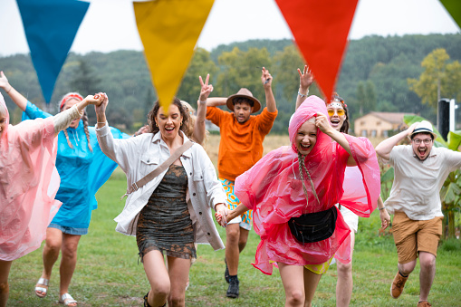 Group of friends of a wide range of ages dressed in waterproof ponchos/festival wear on a rainy summers day. They are having fun, laughing while running for cover in a barn from the rain in the south of France. Some of them have their arms in the air and are holding hands.