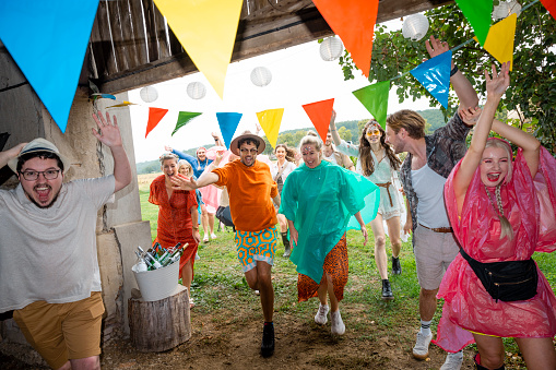 Group of friends of a wide range of ages dressed in waterproof ponchos/festival wear on a rainy summers day. They are having fun, laughing while running for cover in a barn from the rain in the south of France. Some of them have their arms in the air.