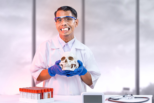 Asian nerd scientist standing and holding a skull head with a stethoscope and medical tube rack on the desk in the laboratory