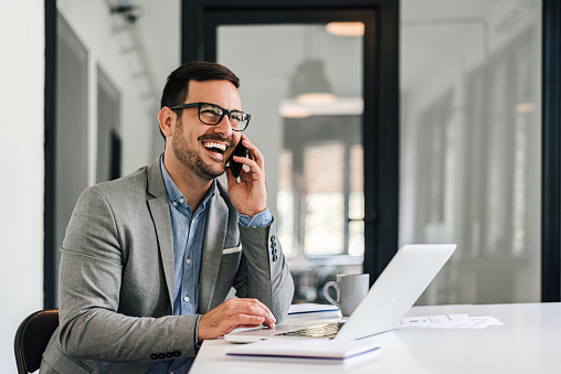 Cheerful businessman wearing formals talking on mobile phone while sitting at office desk