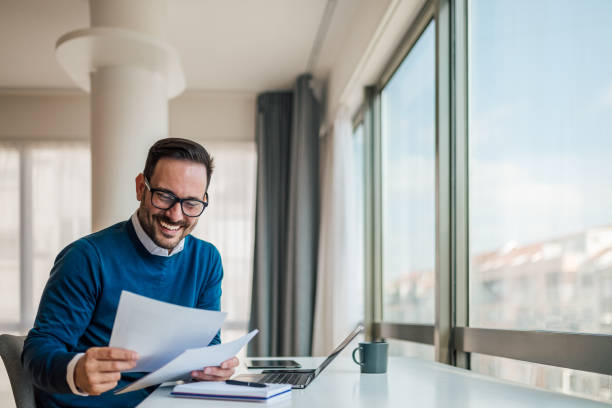 glücklicher geschäftsmann, der dokumente analysiert, während er am schreibtisch im büro sitzt - financial advisor cheerful one person adults only stock-fotos und bilder