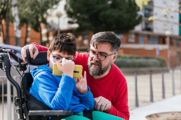 Disabled child in wheelchair watching something on the mobile phone with his father. Disabled child in wheelchair watching something on the mobile phone with his father. Technology and disabled people concept. disabled sign stock pictures, royalty-free photos & images