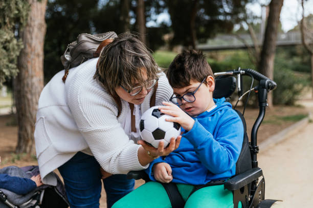 kind mit mehrfachbehinderung im rollstuhl, das mit seiner mutter im freien mit einem fußball spielt. - disabled accessible boarding sign stock-fotos und bilder