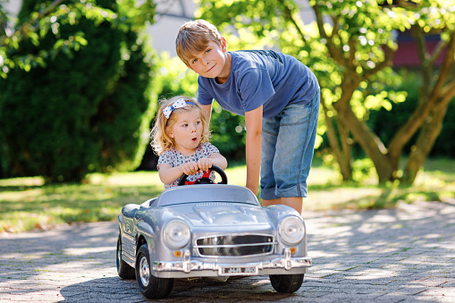 Two happy children playing with big old toy car in summer garden, outdoors. Boy driving car with little girl, cute sister inside. Laughing and smiling kids. Family, childhood, lifestyle concept