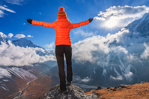 Young woman, wearing orange jacket, lifts her arms in victory. She is standing on the top of a mountain and watching sunset over Himalayas .Mount Everest National Park. This is the highest national park in the world, with the entire park located above 3,000 m ( 9,700 ft). This park includes three peaks higher than 8,000 m, including Mt Everest.