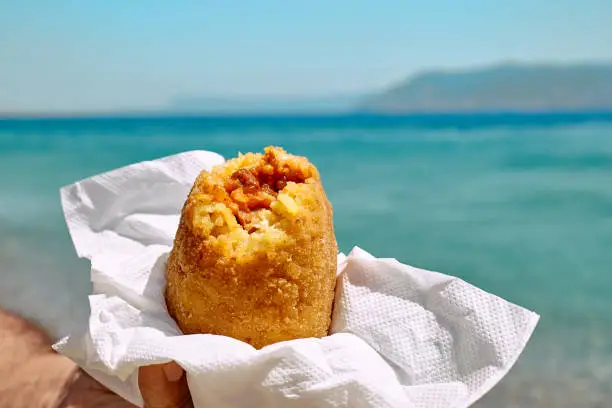 Typical Sicilian street food on seaside background. Hand of man holding hot palatable arancina (deep fried rice balls with meat) in the beach. Take out food.